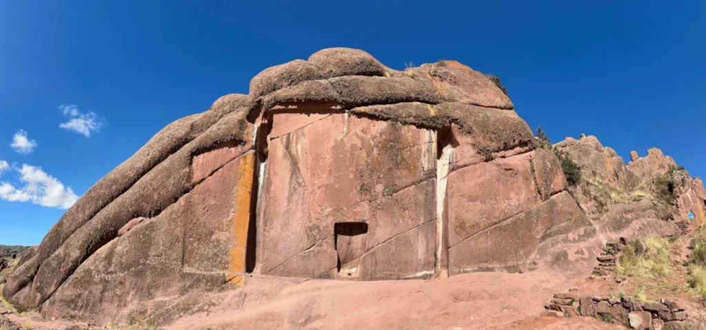 Wide-angle view of the aramu muru interdimensional portal carved into a reddish rock wall under a clear blue sky, with surrounding natural rock formations and a small path leading up to the site.