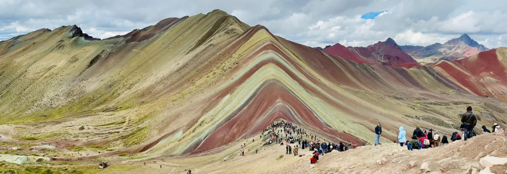 Vinicunca Rainbow Mountain colourful ridge near Cusco with hikers gathered along the viewpoint trail under a cloudy Andean sky.